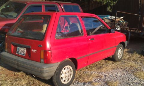 1991 Ford Festiva - A red 1991 Ford Festiva is parked outdoors next to a red truck, with a trailer in the background.