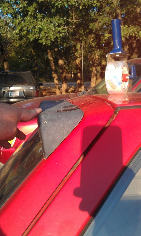 1991 Ford Festiva - A hand points to black trim on a red car's hood, with another vehicle visible in the background.