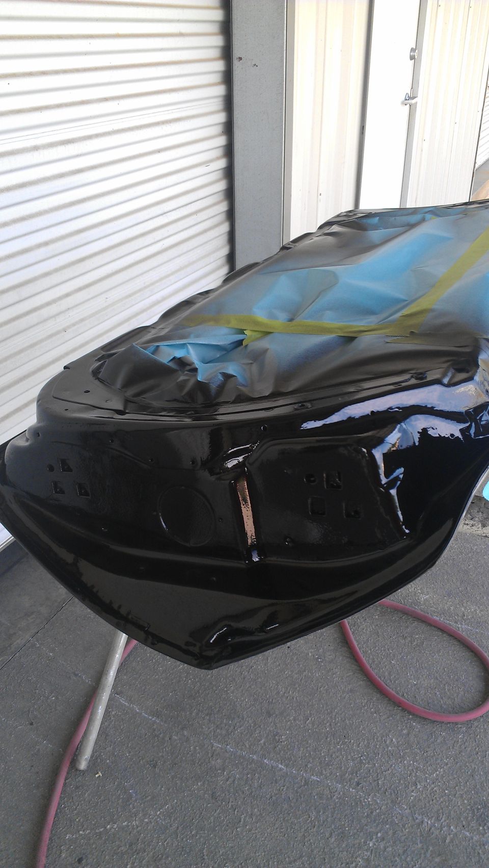 Black car hood covered in plastic sheeting and tape, undergoing painting