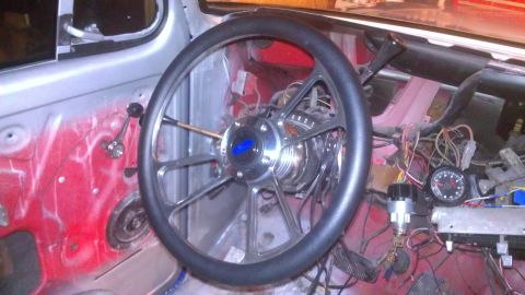 1978 Ford Bronco - The interior of a vehicle is shown, a custom steering wheel with a blue Ford emblem and a dashboard with exposed wiring and a gauge.