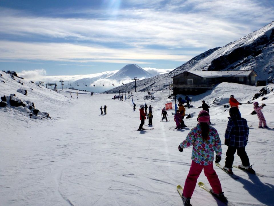 Skiers on a snowy mountain slope with a lodge and a distant volcano under a cloudy sky