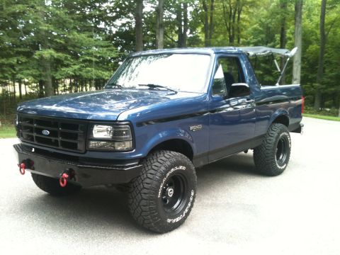 1996 Ford Bronco - A blue Ford Bronco with a custom bumper and large tires sits on a paved surface with trees in the background.