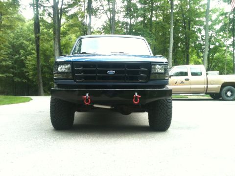 1996 Ford Bronco - A dark blue Ford truck is parked in front of a tan pickup truck on a paved surface with trees in the background.