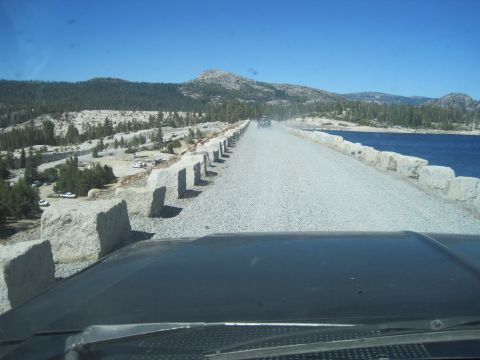 1989 Ford Bronco - A vehicle drives on a gravel road next to a lake, with other vehicles parked on the left side of the road.