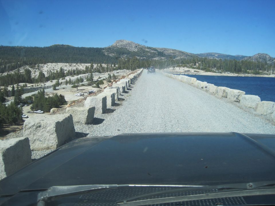 Black dashboard and windshield of a vehicle looking out at a gravel road with a blue lake to the right and mountains in the distance