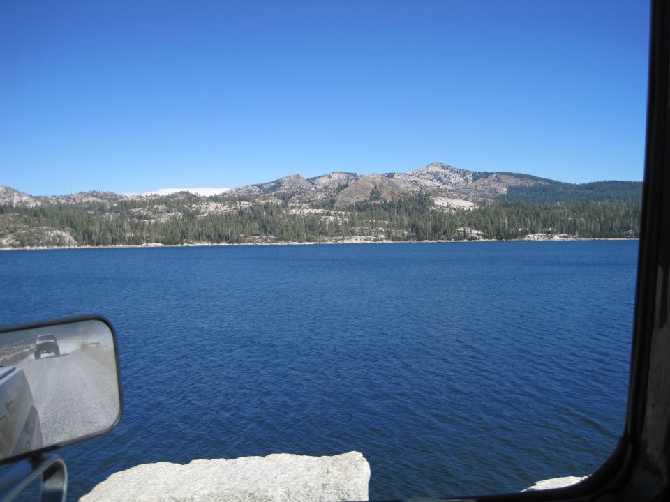 View of a blue lake with tree-covered mountains in the background, seen from inside a vehicle