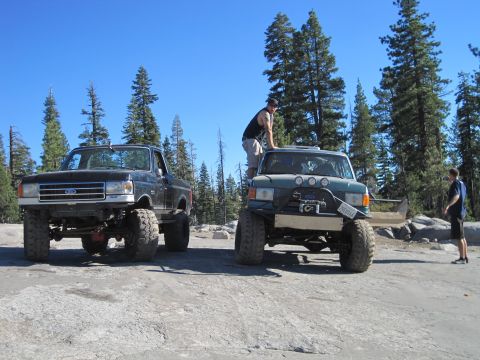 1989 Ford Bronco - Two modified trucks are parked on a rocky surface under a clear blue sky, with a forest in the background.