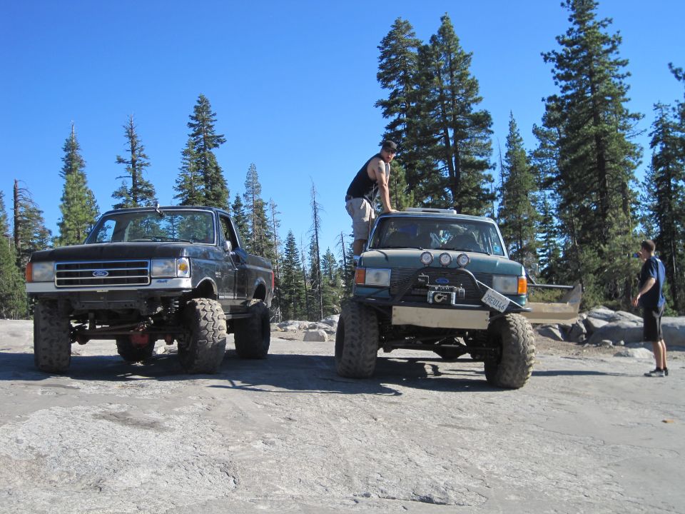 Black Ford truck and a green custom off-road vehicle parked on rocky ground with trees behind them