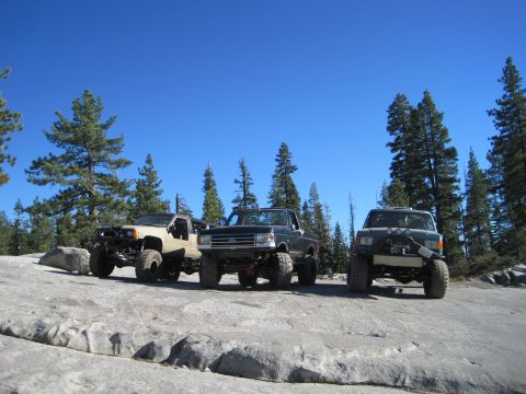 1989 Ford Bronco - Modified trucks are parked on a rocky terrain under a clear blue sky, with pine trees surrounding the area.