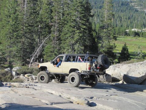 1989 Ford Bronco - Modified SUV navigates a challenging rocky trail surrounded by dense forest and mountainous landscape.
