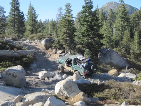 1989 Ford Bronco - This modified SUV is tackling a challenging off-road path surrounded by trees and large boulders.