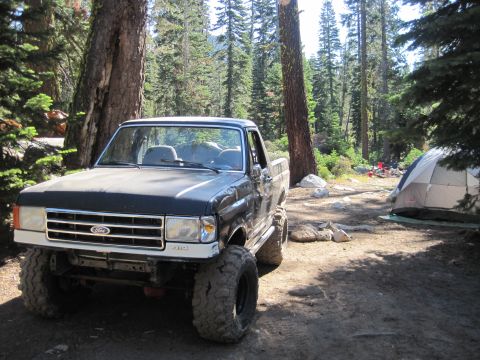 1989 Ford Bronco - A lifted Ford pickup truck is parked in a wooded campsite with a tent set up nearby.