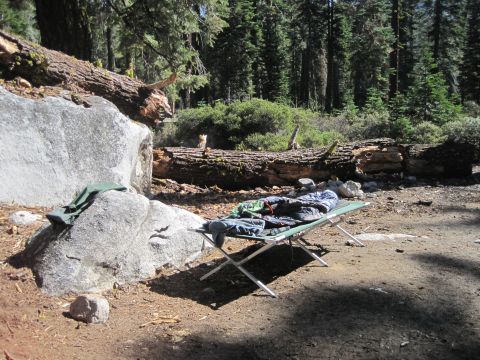 1989 Ford Bronco - A campsite is set up with cots, sleeping bags, and clothes near large rocks and fallen trees in a forest.