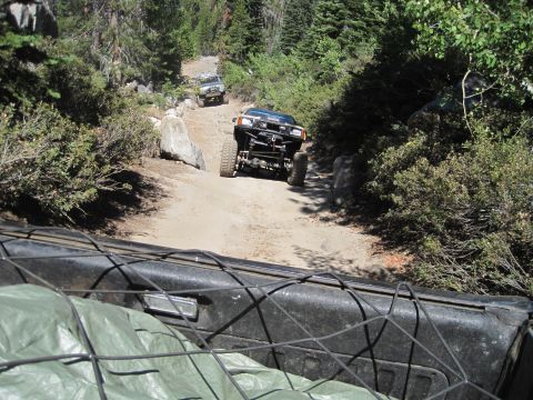 1989 Ford Bronco - Multiple off-road vehicles navigate a challenging, rocky trail through a dense forest on a sunny day.