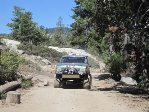 1989 Ford Bronco - This teal SUV, likely a 1987 model, navigates a dusty dirt road with off-road modifications.