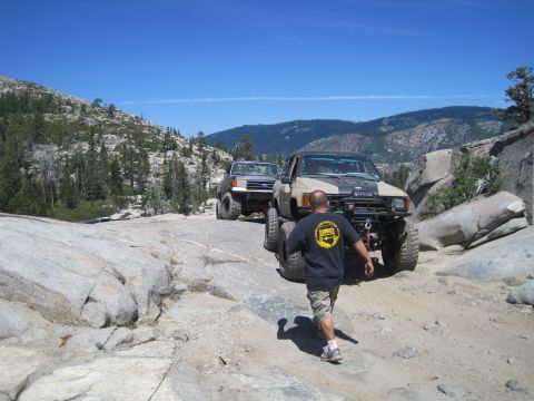 1989 Ford Bronco - Two modified off-road vehicles navigate a challenging rocky path under a clear blue sky.