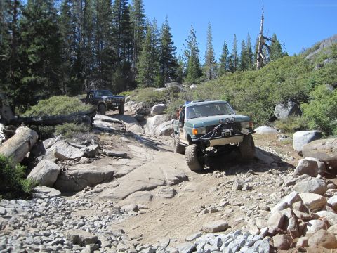 1989 Ford Bronco - Two modified off-road vehicles tackle a challenging, boulder-strewn path in a wooded, mountainous area.