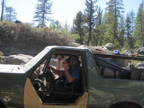 1989 Ford Bronco - Two men drive a green Ford Bronco packed with gear through a wooded, rocky landscape.