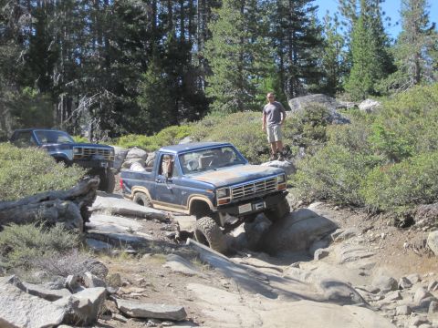 1989 Ford Bronco - Two blue Broncos tackle a challenging rocky trail in a wooded area.