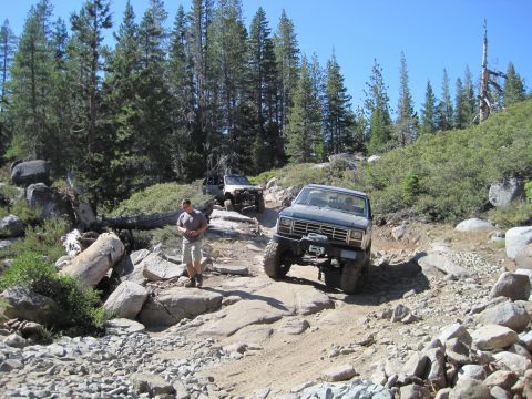 1989 Ford Bronco - A blue SUV with a winch navigates a challenging, rocky trail with two other off-road vehicles following in the background.