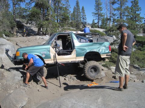 1989 Ford Bronco - A man works on the front tire of a modified Ford Bronco on a , rocky terrain.