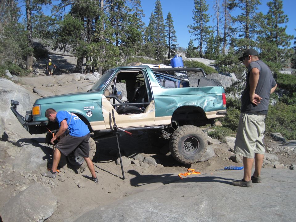 Green Ford Bronco with tan lower body panels, off-roading on a rocky trail with two men nearby