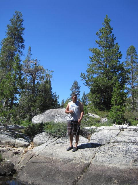 1989 Ford Bronco - A man stands on a rocky outcrop in a natural setting with trees and a bright blue sky overhead.