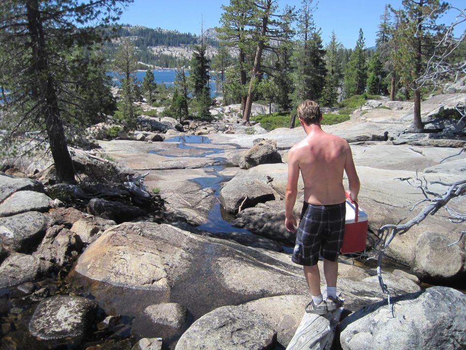 Man with plaid shorts and a red cooler walks over rocks near a stream, with a lake and trees in the background