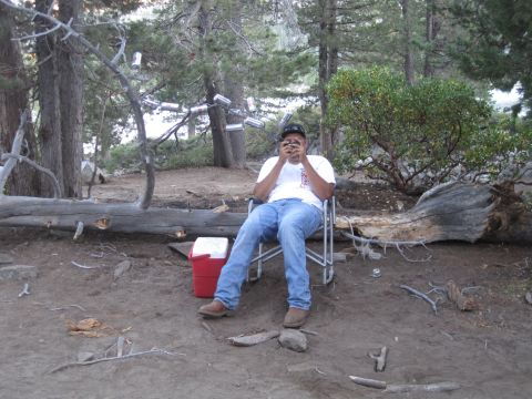 1989 Ford Bronco - A person relaxes in a camping chair outdoors, with a cooler nearby and cans hanging from a tree branch.