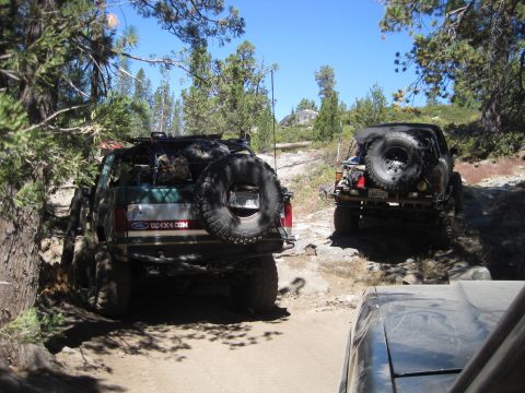 1989 Ford Bronco - Two SUVs tackle a challenging off-road path surrounded by trees and rocks under a clear blue sky.