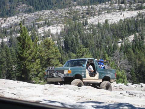 1989 Ford Bronco - A modified green SUV navigates a challenging rocky terrain with a backdrop of pine trees and mountains.