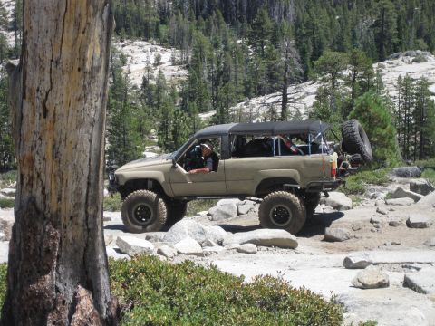1989 Ford Bronco - A tan off-road SUV navigates a rocky trail, its lifted suspension and soft top configuration.