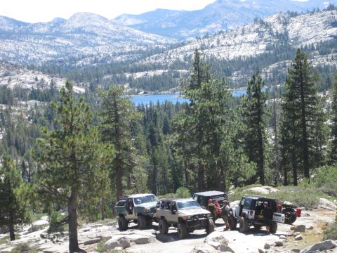 1989 Ford Bronco - Off-road vehicles are parked on a rocky trail with a scenic mountain and lake backdrop.