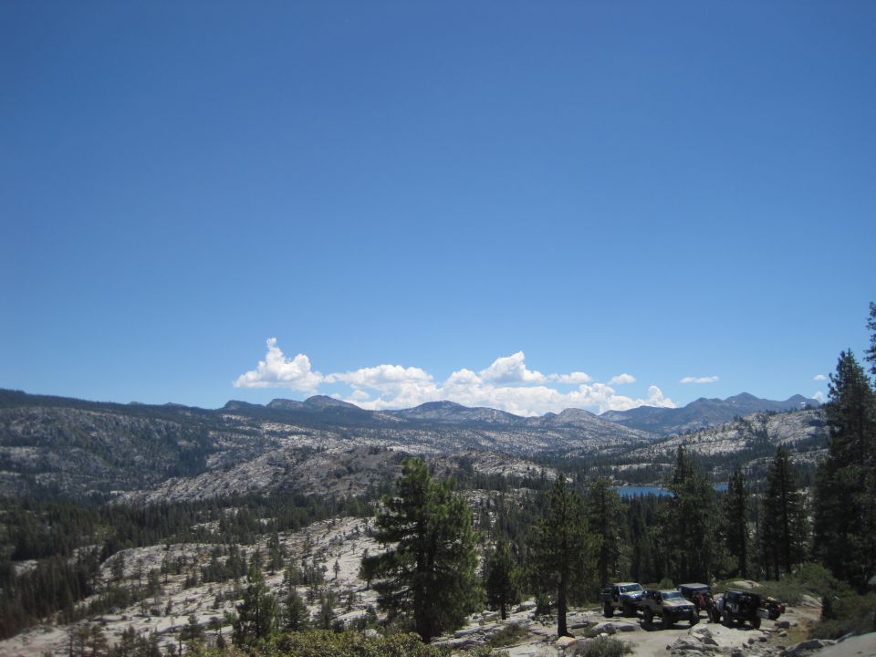 Four SUVs parked on a rocky, wooded slope with a distant lake and mountains