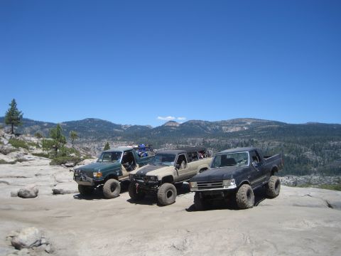 1989 Ford Bronco - Three trucks are parked on a rocky terrain with a scenic mountain view under a clear blue sky.