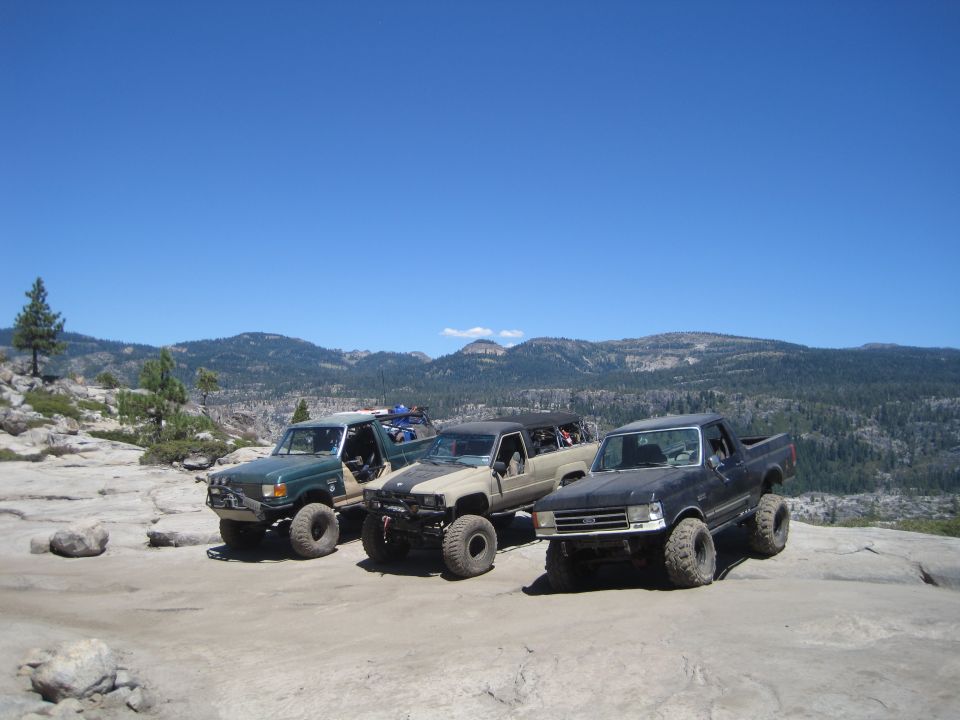 Three modified off-road trucks parked on a rocky overlook with mountains in the background