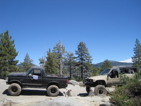 1989 Ford Bronco - Two modified SUVs, a 1989 Ford Bronco and a Toyota 4Runner, are parked on a rocky terrain under a clear blue sky.