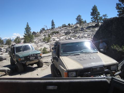 1989 Ford Bronco - Two modified trucks are shown tackling a challenging, rocky off-road path on a sunny day.