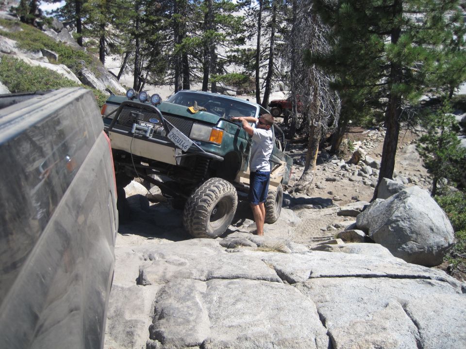 Green off-road SUV with large tires and a winch, navigating a rocky trail. A person is leaning on the hood