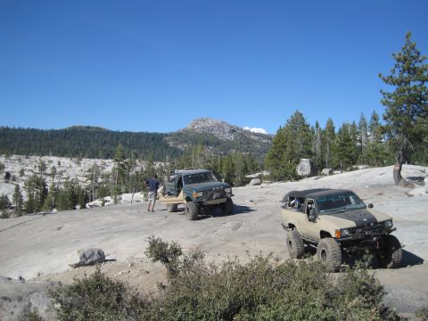1989 Ford Bronco - Two off-road vehicles are parked on a rocky surface with trees and mountains in the background.