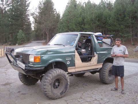 1989 Ford Bronco - A modified green Ford Bronco with off-road tires and a custom bumper stands parked on a dirt road.