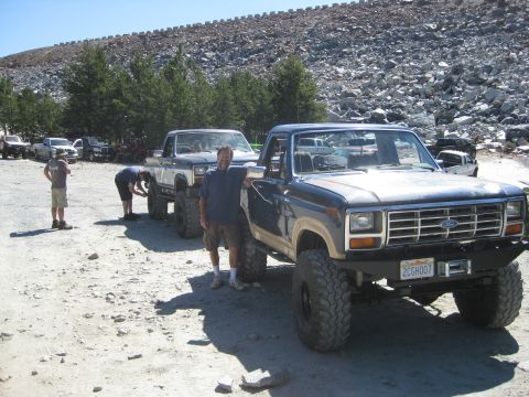 1986 Ford Bronco - Several off-road vehicles are parked on a gravelly surface with a rocky hillside in the background.