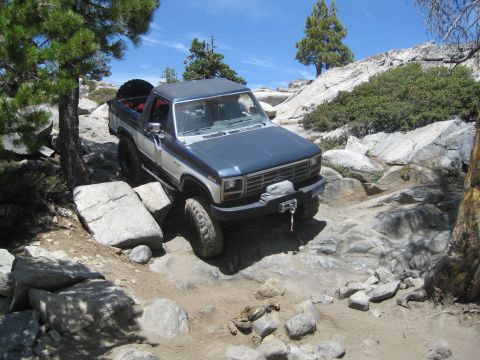 1986 Ford Bronco - This SUV is tackling a challenging off-road path with large rocks and uneven terrain.