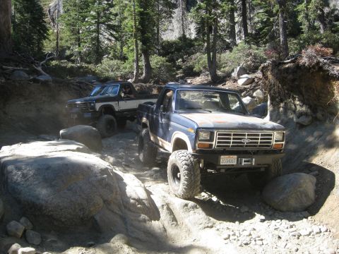 1986 Ford Bronco - Two off-road vehicles navigate a challenging, boulder-strewn path through a wooded area.