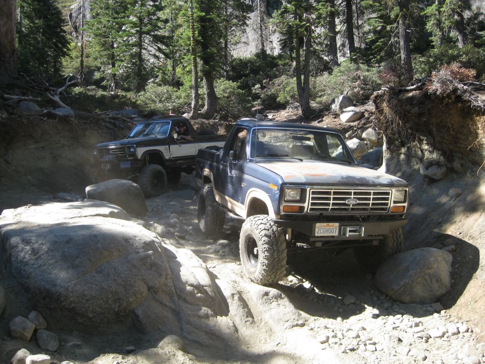 Blue Ford Bronco and a black and white pickup truck driving on a rocky trail in a forest