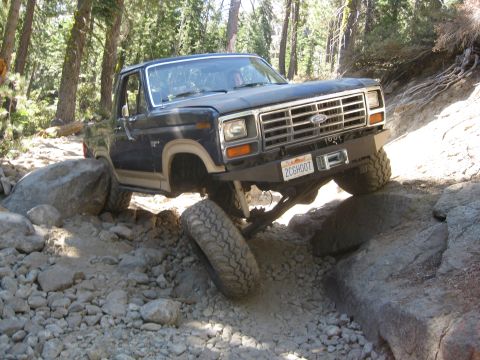 1986 Ford Bronco - This Ford Bronco is shown off-roading on a rocky path with trees in the background.