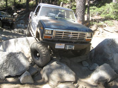 1986 Ford Bronco - A blue Ford truck navigates a challenging rocky trail, with another similar vehicle visible in the background.