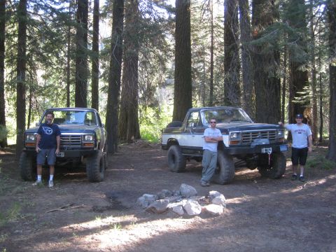1986 Ford Bronco - Two lifted Ford Broncos are parked in a forest clearing with people standing by them.