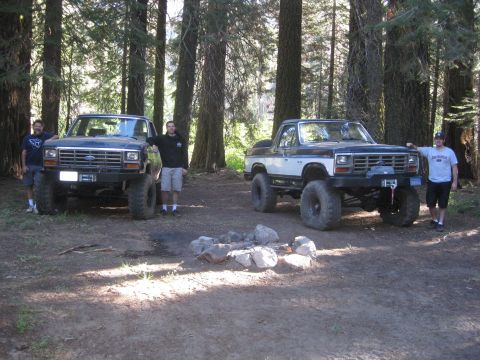 1986 Ford Bronco - Two lifted Ford pickup trucks are parked in a forest with two men standing next to them.