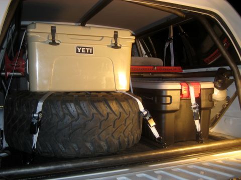 1996 Ford Bronco - The cargo area of a vehicle is organized with a spare tire, a Yeti cooler, and a black storage box secured with straps.